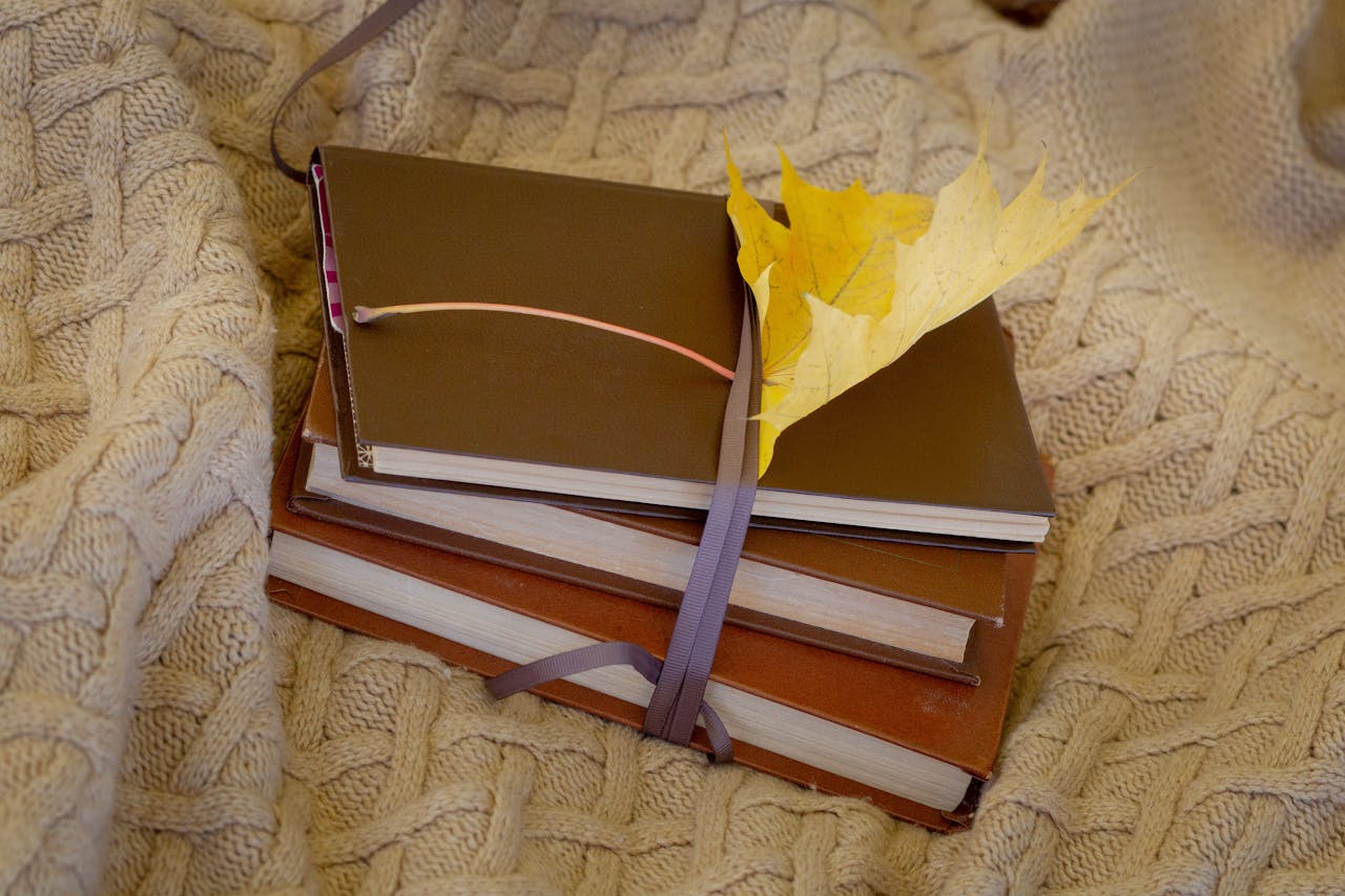 Stack of books with a yellow leaf on a knitted blanket, conveying a warm autumn vibe.
